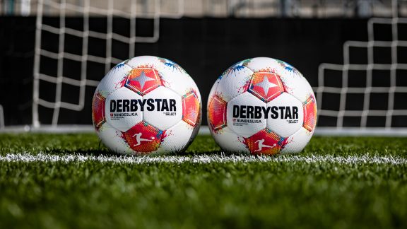 Die offiziellen Spielbälle der Bundesliga und 2. Bundesliga liegen nebeneinander auf einem Fußballfeld. Im Hintergrund ein Tor.