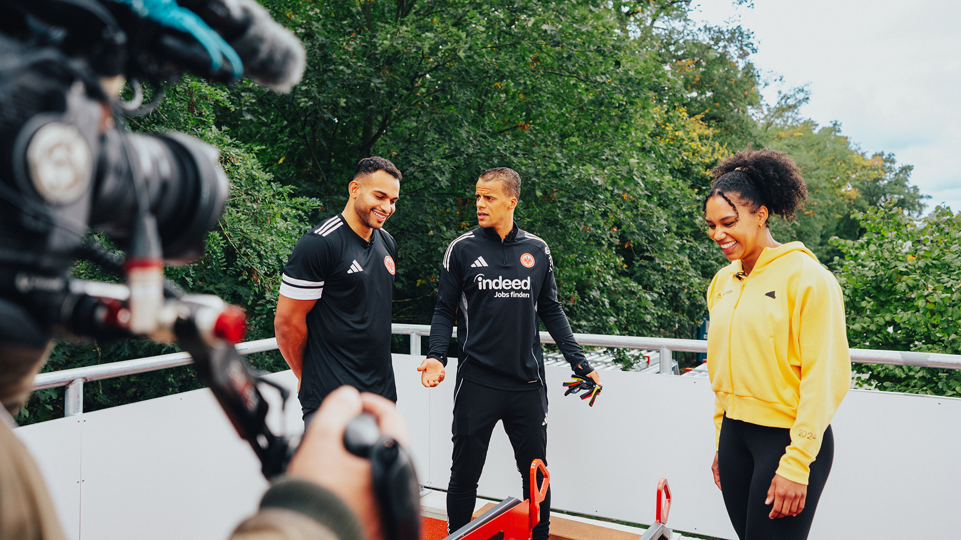 Eine Kamera filmt drei Personen, die auf einer Terrasse oder Plattform im Freien stehen. Zwei von ihnen tragen schwarze Trainingskleidung, die dritte Person trägt eine gelbe Sporthilfe-Jacke und schwarze Leggings. Im Hintergrund sind grüne Bäume zu sehen. Die Szene wirkt wie eine sportliche Interview- oder Trainingssituation.