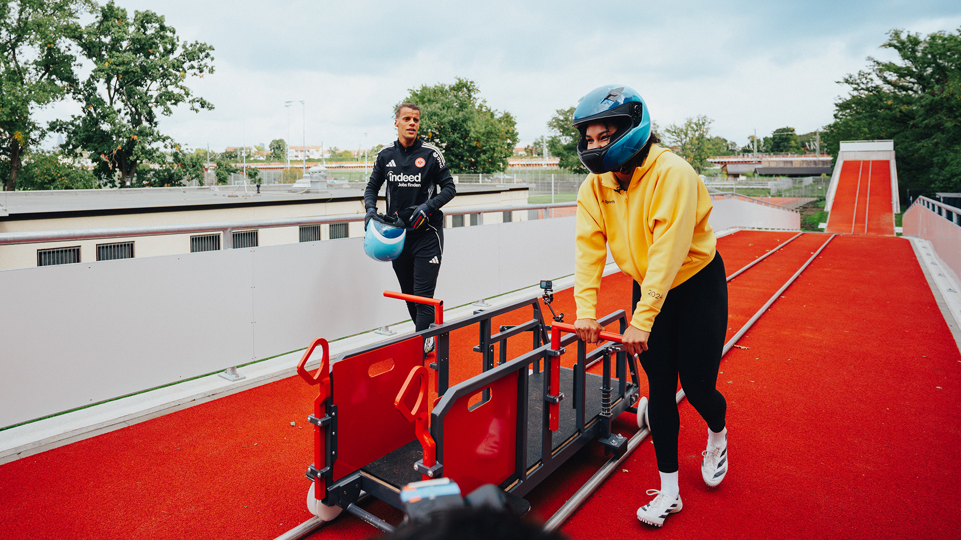 Auf einer roten Bobbahn im Freien steht eine Person in gelber Sportjacke und schwarzen Leggings mit einem blauen Helm und hält einen Bob-Anschubschlitten fest. Eine weitere Person in schwarzer Trainingskleidung und mit einem blauen Helm in der Hand steht im Hintergrund. Die Szene zeigt eine sportliche Trainingssituation im Bereich Bobsport.
