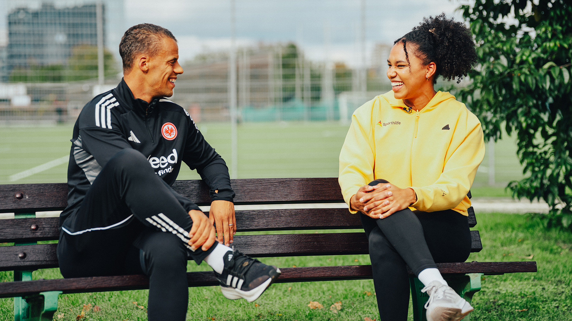 Zwei Personen sitzen auf einer Parkbank im Freien und unterhalten sich. Die Person links trägt einen schwarzen Trainingsanzug, während die Person rechts eine gelbe Sportlife-Jacke und schwarze Leggings trägt. Im Hintergrund ist ein Sportplatz mit grüner Wiese und ein modernes Gebäude zu sehen. Die Szene wirkt entspannt und sportlich.