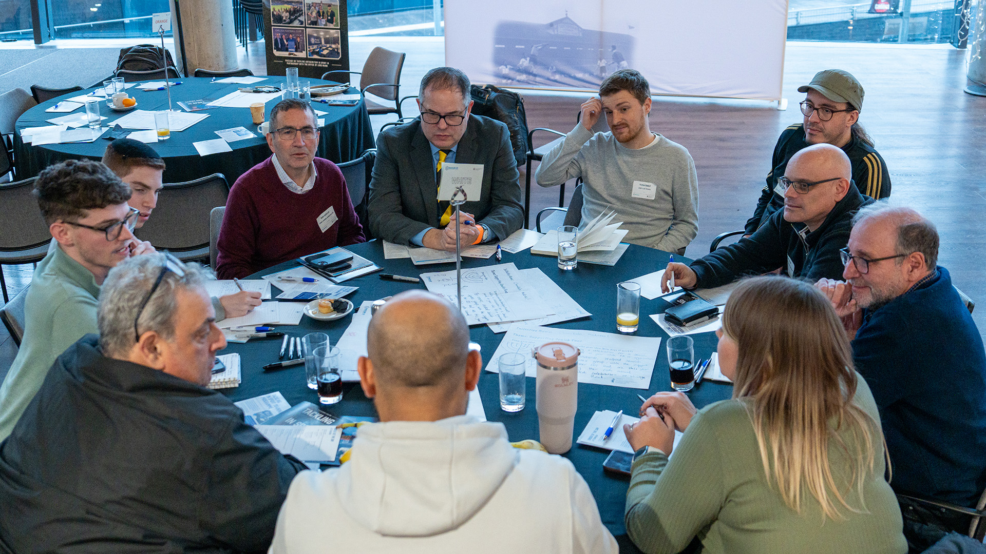 Menschen sitzen gemeinsam an einem runden Tisch und diskutieren bei einem Workshop, auf dem Tisch liegen Notizen und Getränke.