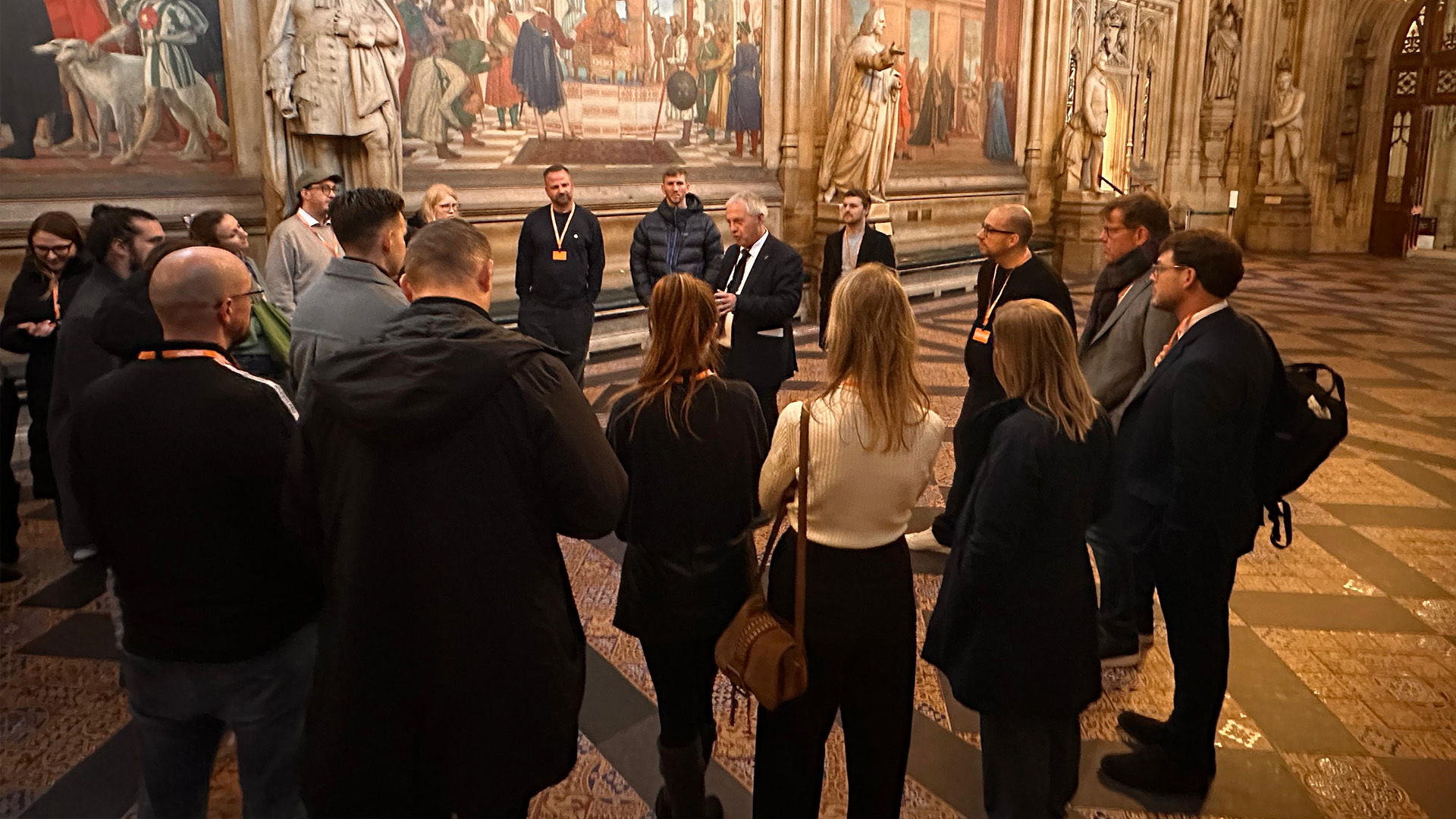 Eine Gruppe von Menschen steht in einer historischen Halle und hört einer Führungsperson zu, im Hintergrund sind große Gemälde und Statuen zu sehen.