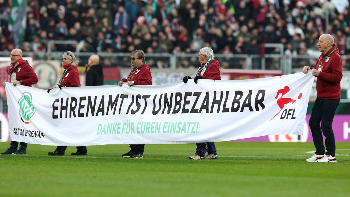 Personen in roten Jacken halten im Stadion ein weißes Banner mit der Aufschrift „Ehrenamt ist unbezahlbar“ und „Danke für euren Einsatz!“, daneben sind die Logos der DFL und „Aktion Ehrenamt“ zu sehen. Im Hintergrund sitzen Zuschauer.