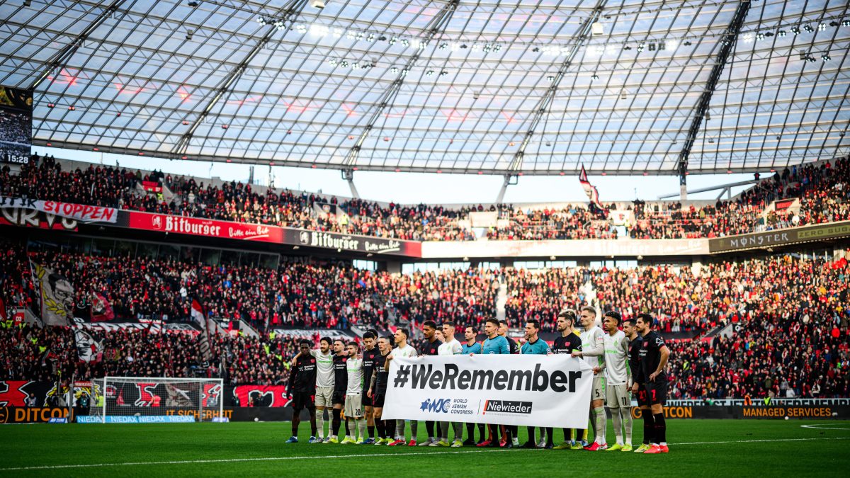 Spieler in schwarzen und weißen Trikots stehen nebeneinander im Stadion und halten gemeinsam ein großes Banner mit der Aufschrift "#WeRemember". Im Hintergrund sind zahlreiche Zuschauer auf den Tribünen zu sehen.