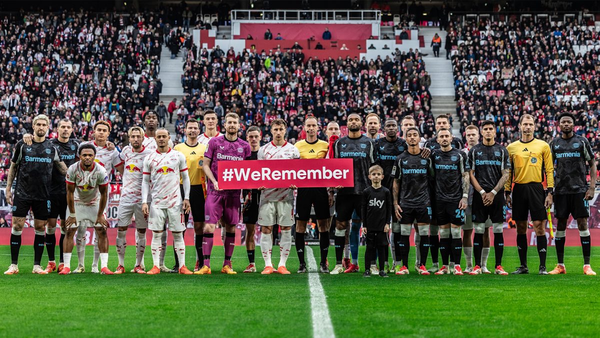 Players from two football teams and referees stand together on the pitch holding a "#WeRemember" sign, with a packed stadium in the background, showing solidarity and remembrance before the match.